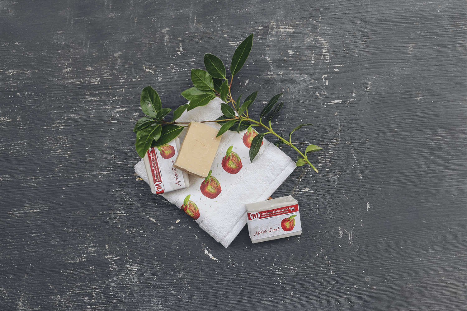 Lanolin-Naturseife mit Apfel-Zimt-Duft, mit grünem Zweig und Gästehandtuch mit Apfel-Motiv.