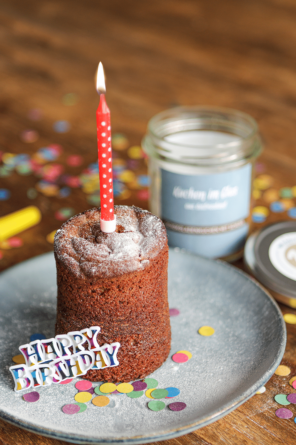 Schokoladenkuchen mit Schuss, auf einem Teller mit Kerze, Konfetti und einem Schriftzug Happy Birthday angerichtet.