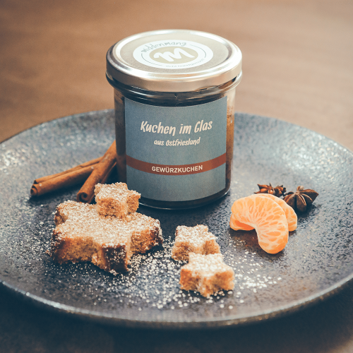 Gewürzkuchen im Glas, in Sternform ausgestanzt auf einem Teller angerichtet, mit Puderzucker, Gewürzen und Mandarinenstücken.