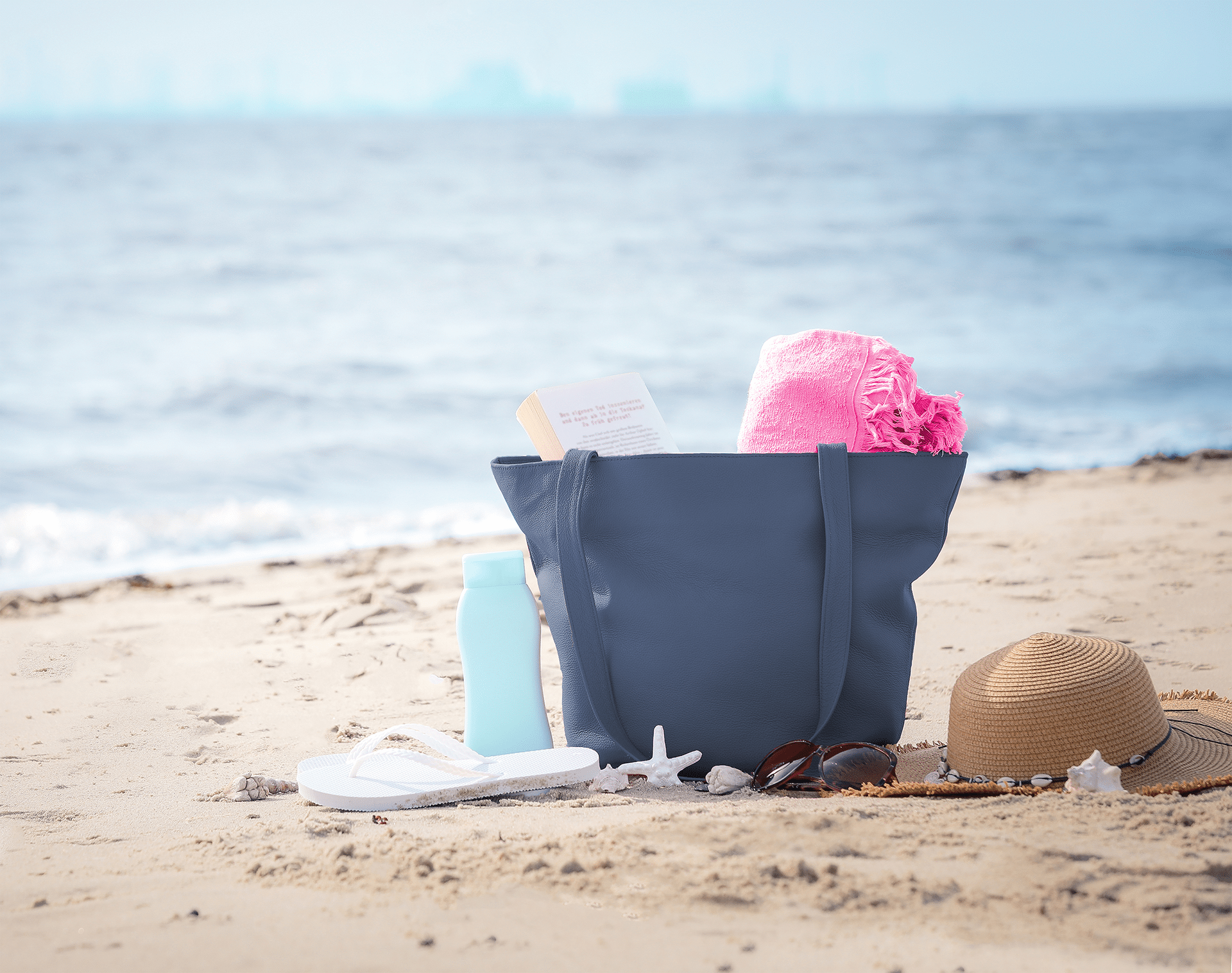 Eine Handtasche aus Leder in der Farbe "atlantic" steht am Strand mit Strandartikeln.