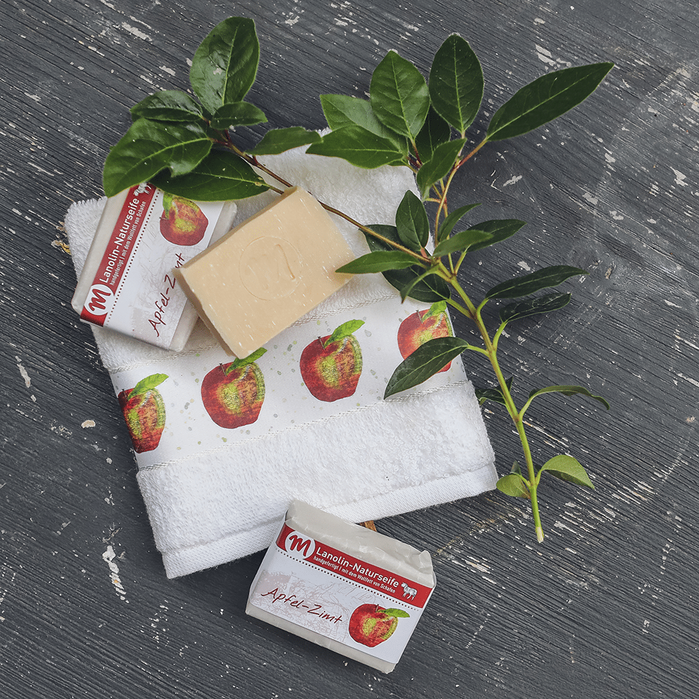 Lanolin-Naturseife mit Apfel-Zimt-Duft, mit grünem Zweig und Gästehandtuch mit Apfel-Motiv.