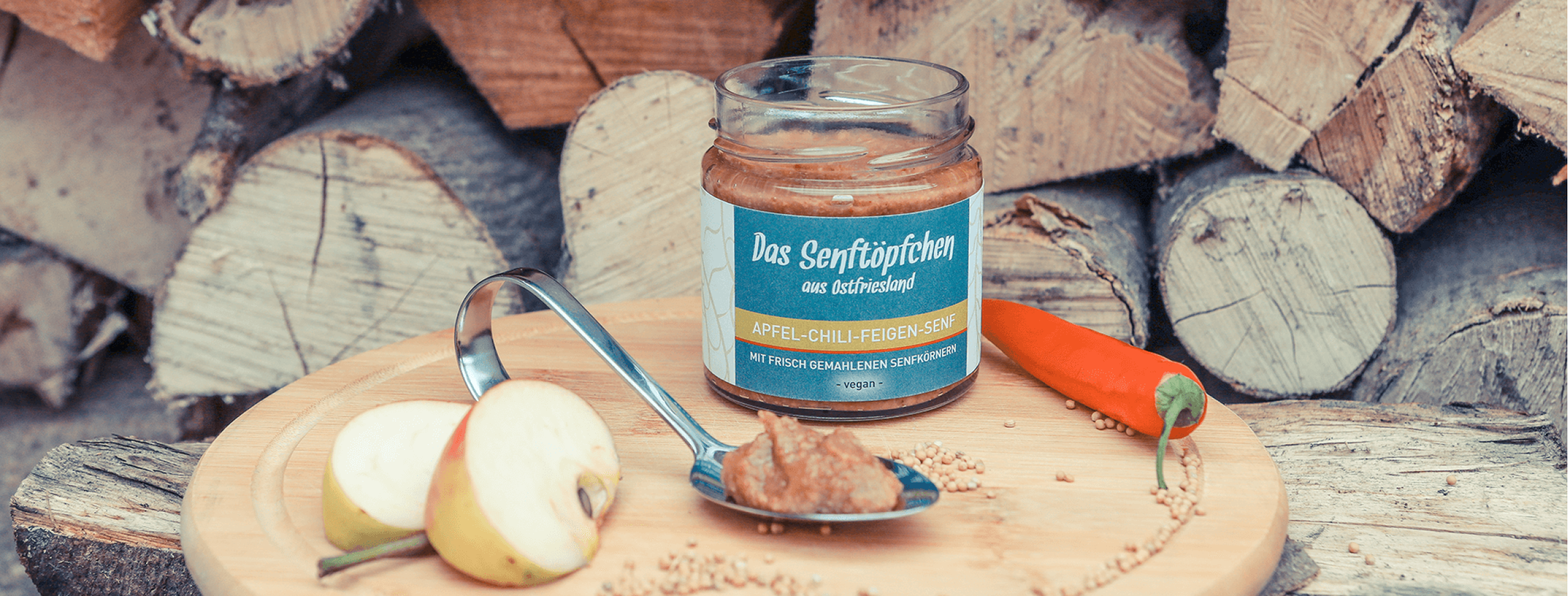 Senftöpfchen Senfglas mit orangenem Inhalt und blauem Etikett auf einem Holzbrett umgeben von einem Löffel, Apfel, Chili und Senfkörnern.