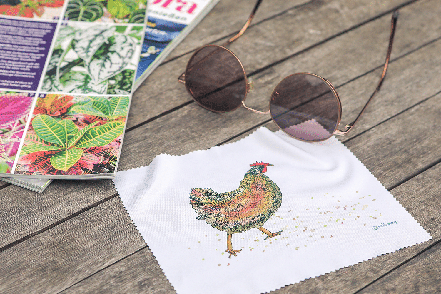 Ein Brillenputztuch mit dem Motiv Huhn liegt vor einer Sonnenbrille.