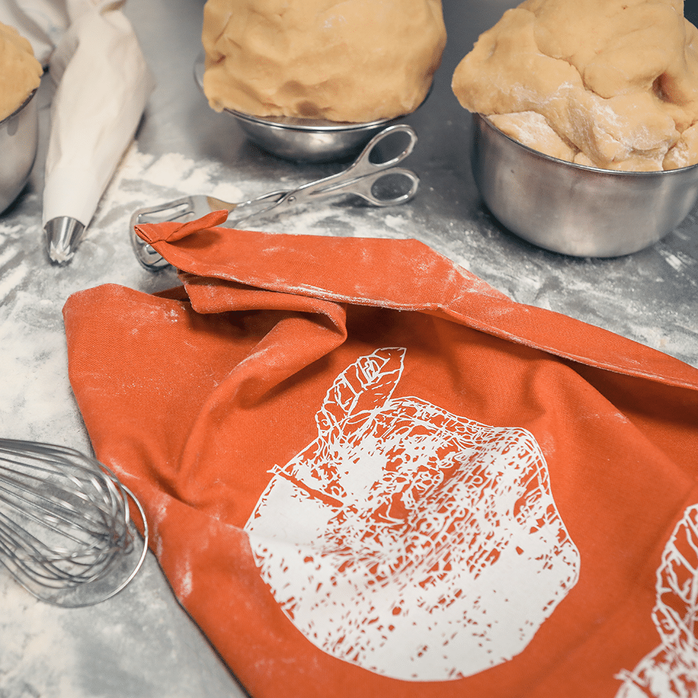 Ein rost-oranges Geschirrtuch bedruckt mit roten Äpfeln, auf einer Anrichte mit Mehl und Teig.