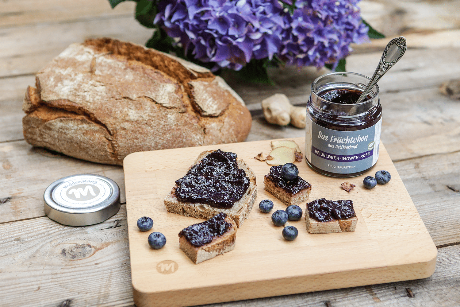 Der geöffnete Fruchtaufstrich Heidelbeer-Ingwer-Rose steht auf einem Holzbrett, daneben liegt Brot mit dem Aufstrich.