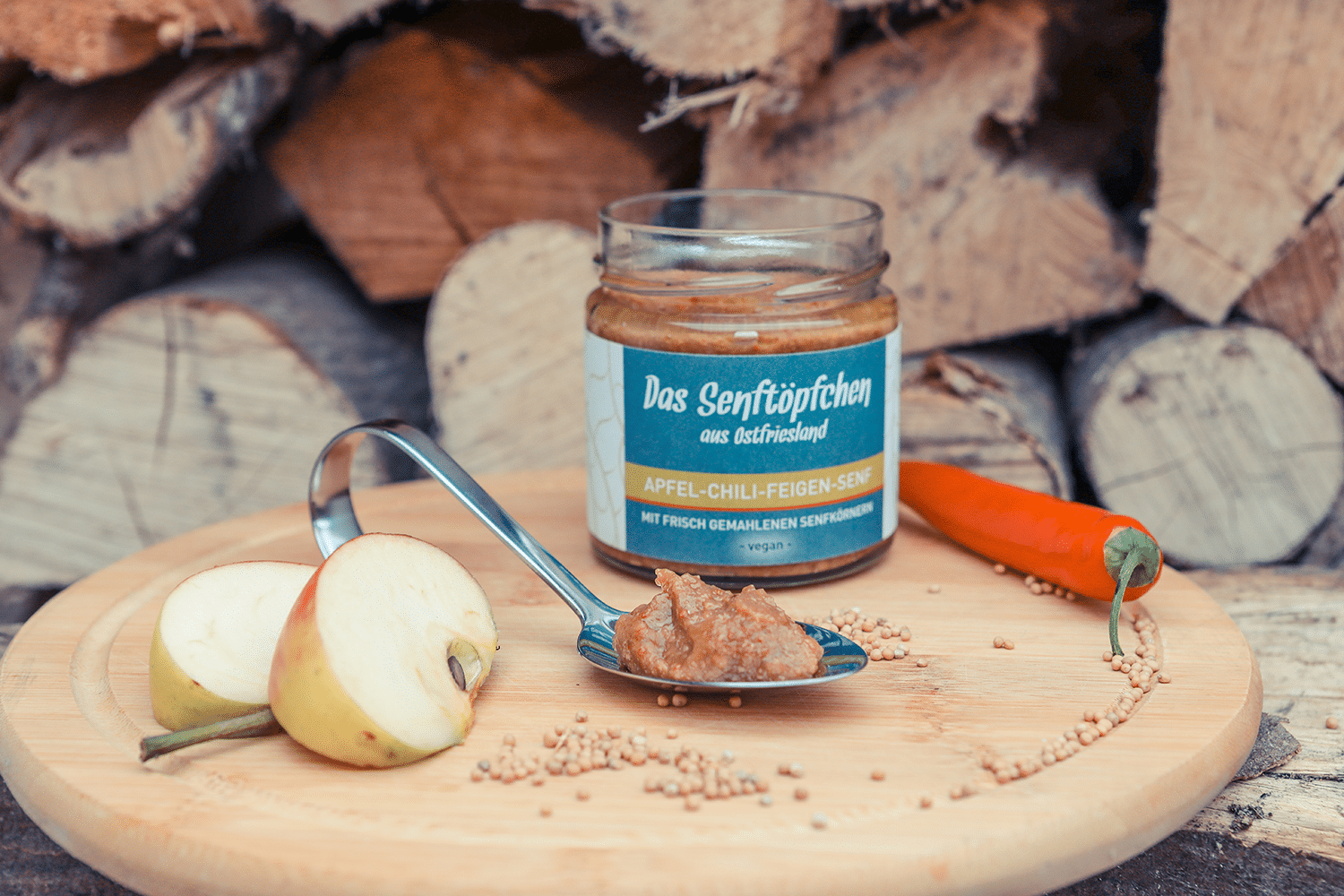 Ein geöffnetes Glas Apfel-Chili-Feigen-Senf auf einem Holzbrett mit einem gefüllten Anrichtelöffel.