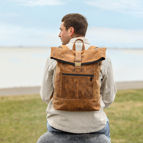 Person trägt den Leder-Rucksack in hellbraun auf dem Rücken und sitzt am Deich.