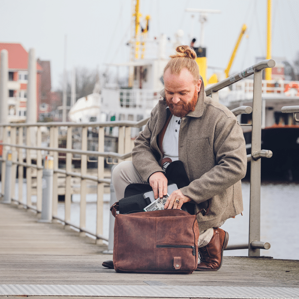 Hockende Person holt sich ein Buch aus der Leder-Umhängetasche in braun.