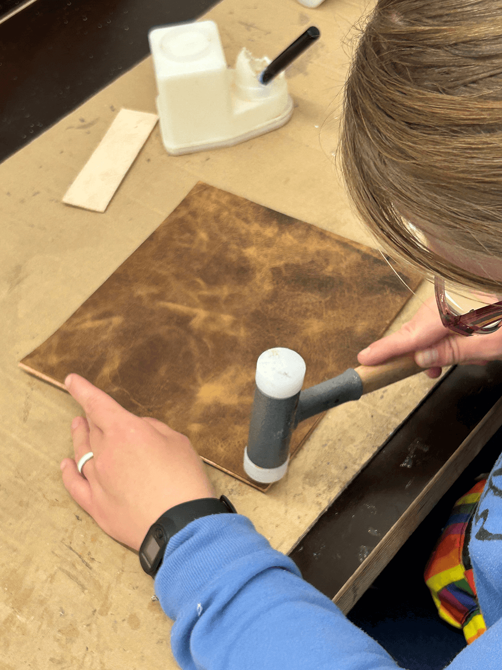 Eine Person bearbeitet mit einem Gummihammer ein Stück hellbraunes Leder auf einer Werkbank.