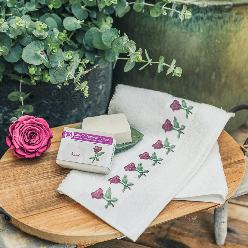 Ein G&auml;stehandtuch auf einem Holztablett, mit Rosen in Altrosa und Gr&uuml;n bedruckt und mit zwei St&uuml;cken Rosenseife daneben.