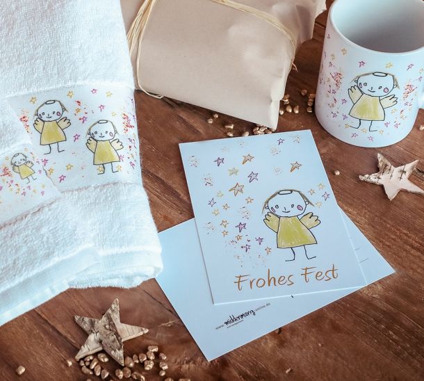Weißes Handtuch, weiße Postkarten und weiße Tasse jeweils mit gelbem Engelsmotiv liegen auf einem Holztisch umgeben von Sternendeko.