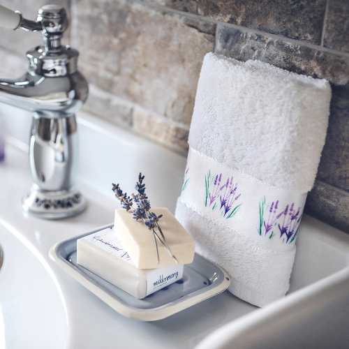 Ein Gästehandtuch, bedruckt mit Lavendel, gefaltet und aufgestellt auf einem Waschbeckenrand, mit einem Stück Seife davor.