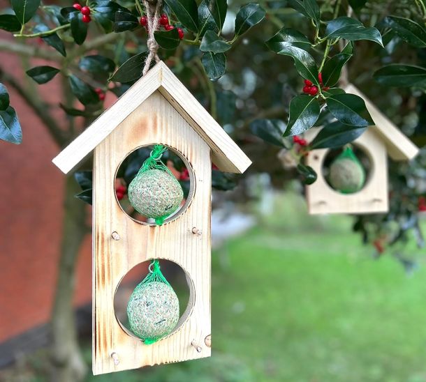 Zwei am Baum hängende Vogelfutterhäuser aus Holz, einmal mit zwei Ein Vogelfutterhaus aus Holz hängt am Baum mit zwei untereinander befestigten Meisenknödeln.