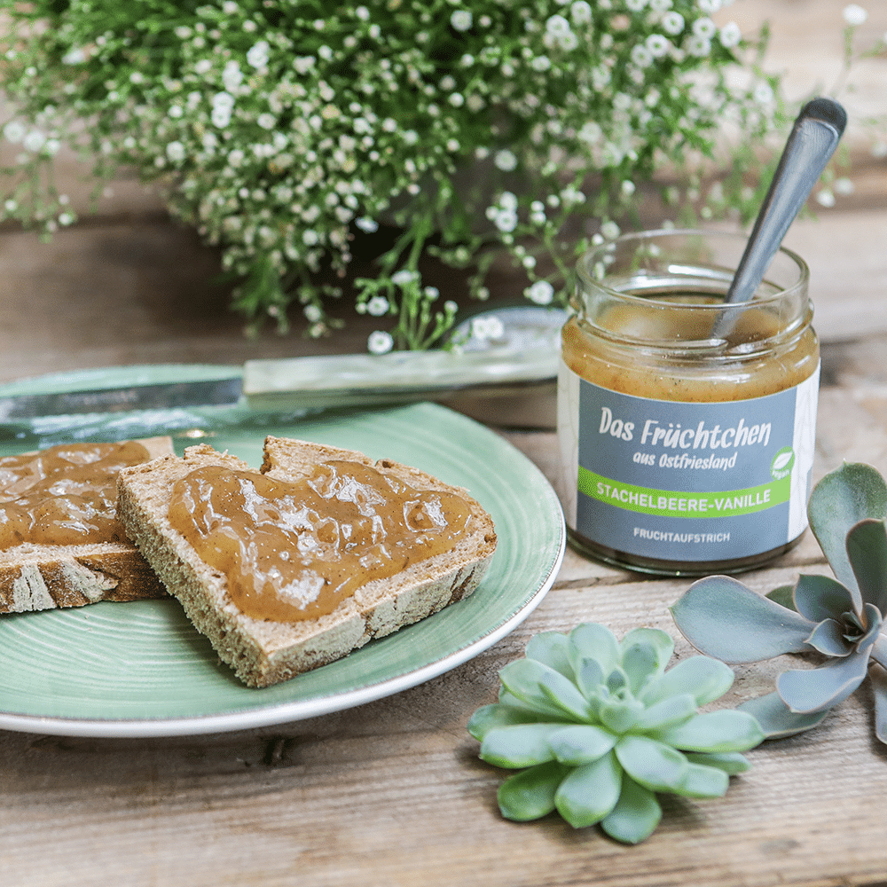 Brotscheiben auf einem Teller, bestrichen mit Stachelbeere-Vanille-Fruchtaufstrich und einem offenen Glas daneben.