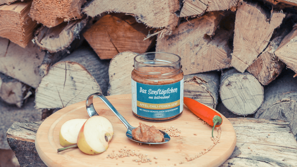 Ein geöffnetes Glas Apfel-Chili-Feigen-Senf auf einem runden Holzbrett, dekoriert mit Chili und Apfelstücken.