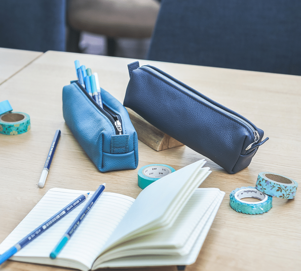 Zwei blaue Federmäppchen aus Leder, mit Stiften und einem aufgeschlagenen Schreibblock.