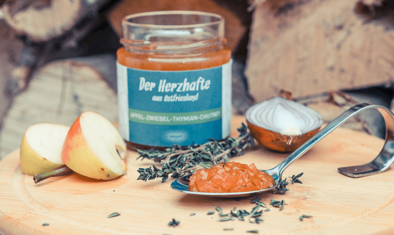 Apfel-Zwiebel-Thymian-Chutney dekoriert mit Apfelspalten, Zwiebelhälfte und Thymianzweig auf Holzbrett.