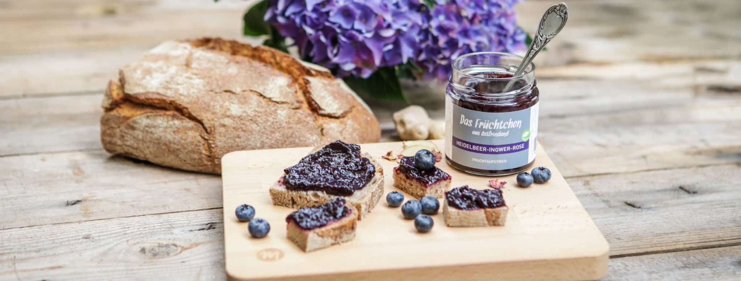 Kleingeschnittenes Brot auf einem Teller, bestrichen mit Heidelbeer-Ingwer-Rose-Fruchtaufstrich und einem offenen Glas daneben.