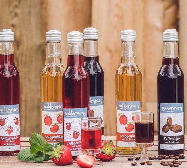 Sechs Flaschen mit verschiedenen Likören auf einem Tisch mit zwei Gläsern und frischen Erdbeeren, sowie Kaffeebohnen.