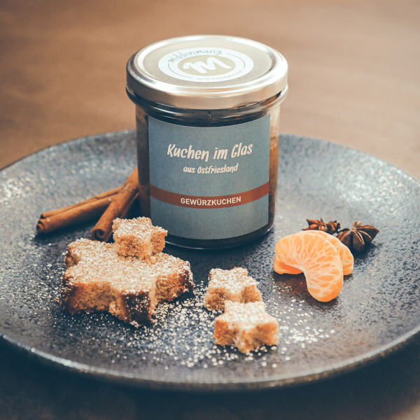 Gewürzkuchen im Glas, in Sternform ausgestanzt auf einem Teller angerichtet, mit Puderzucker, Gewürzen und Mandarinenstücken.