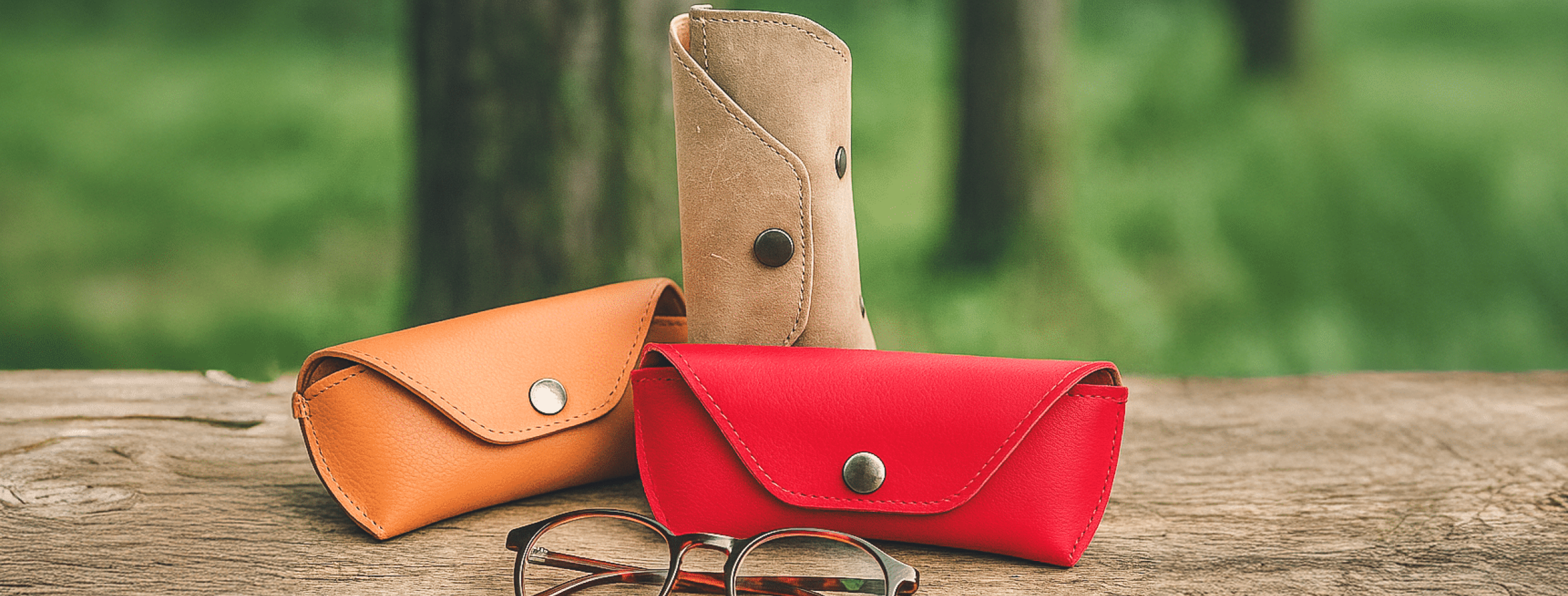 Brillenetuis "Sichtbare" in Herbstfarben arrangiert vor einem waldigen unscharfem Hintergrund, vor den Etuis liegt eine Brille