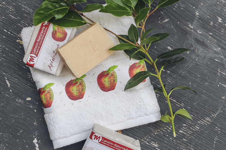 Lanolin-Naturseife mit Apfel-Zimt-Duft, mit grünem Zweig und Gästehandtuch mit Apfel-Motiv.