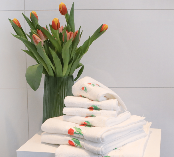 Ein Stapel Handtücher und Gästehandtücher mit Tulpenmotiv, auf einem Hocker, mit einer Vase Tulpen daneben.