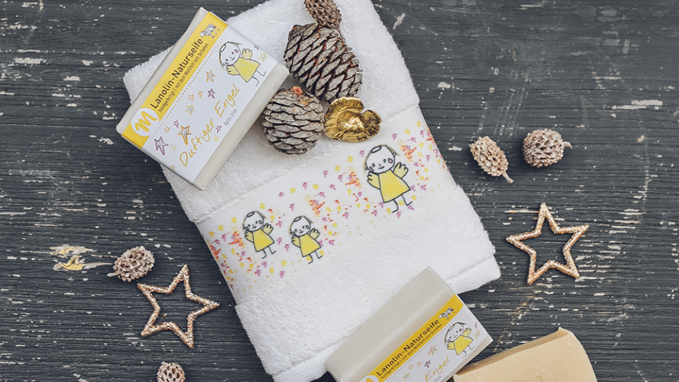 Lanolin-Naturseife mit Apfel-Zimt-Duft und einer Banderole mit duftigem Engel auf einem G&auml;stehandtuch mit Engel-Motiv.