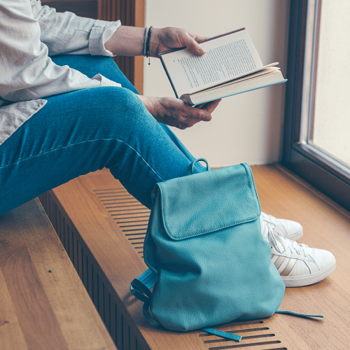 Kleiner Leder-Rucksack in der Farbe rauchblau steht neben einer Person, die in einem Buch liest.