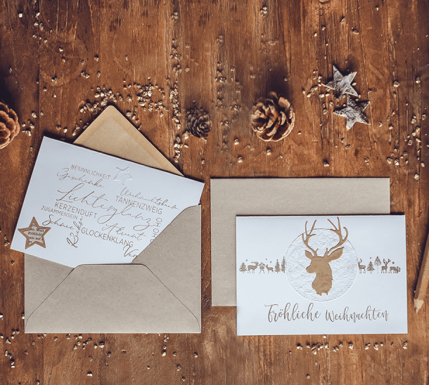 Zwei weiße Klappkarten mit weihnachtlichen Motiven und zwei Umschlägen, weihnachtlich dekoriert.