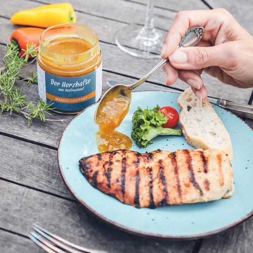 Geöffnetes Glas Mango-Chutney auf einem Teller mit Grillfleisch und Beilagen.