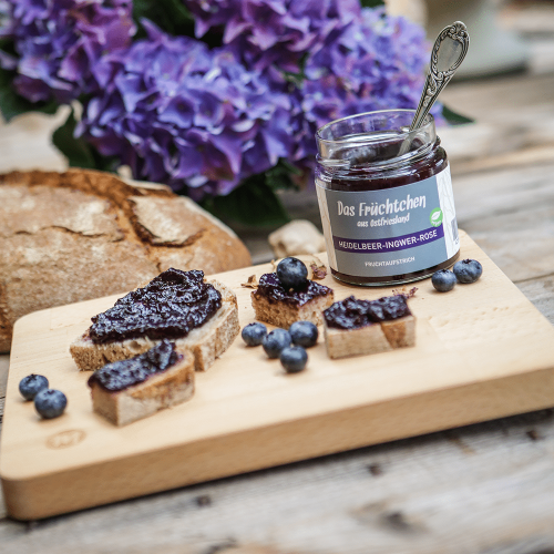 Kleingeschnittenes Brot auf einem Teller, bestrichen mit Heidelbeer-Ingwer-Rose-Fruchtaufstrich und einem offenen Glas daneben.