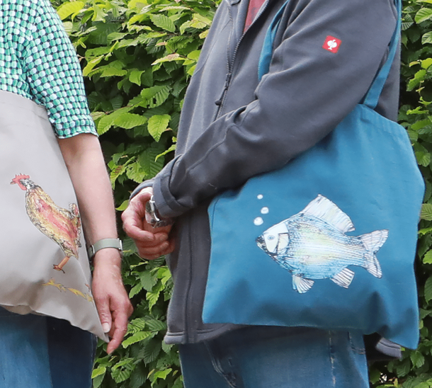 Zwei Personen tragen Stofftaschen über der Schulter. Die Taschen zeigen die Motive Fisch und Huhn.