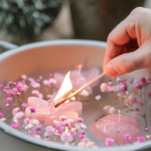 Drei Schwimmkerzen in Form von Biene, K&auml;fer und Libelle, in der Farbe Skin rose, schwimmend in einer Schale mit Bl&uuml;ten. Eine Person entz&uuml;ndet eine Kerze.