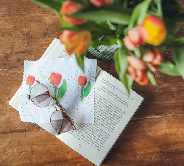 Brillenputztuch mit Motiv Tulpen, mit Lesebrille auf einem offenen Buch mit Tulpenstrauß im Hintergrund.